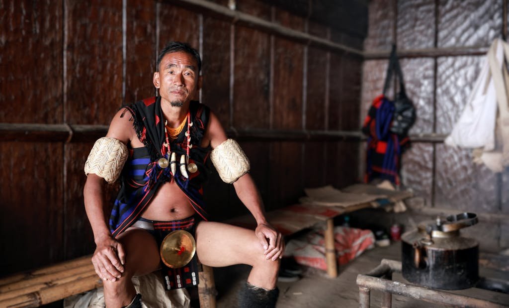 A man in traditional tribal attire from Nagaland, India, sits in a rustic interior setting.